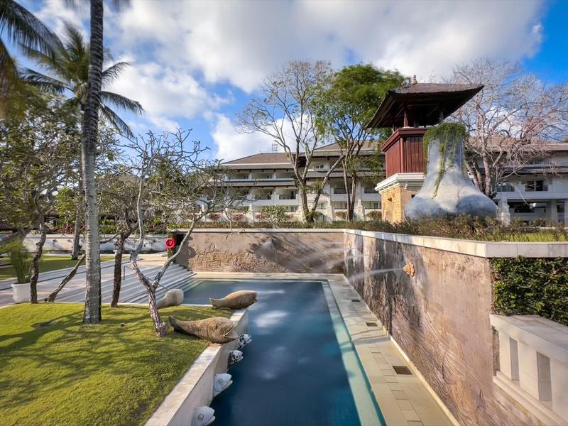 Modern hotel complex with pool, palm trees, and partly cloudy sky