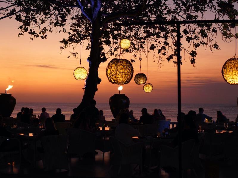 Atmospheric beach bar at sunset with hanging lights and guests by the sea.