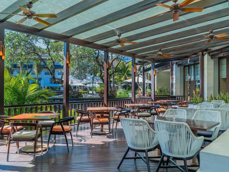 Covered terrace with modern seating and fans, surrounded by greenery at a hotel.