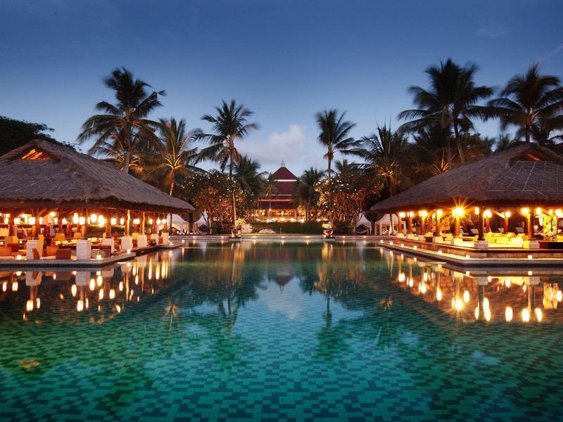 Lit resort with pool, surrounded by palm trees and tropical buildings at dusk.