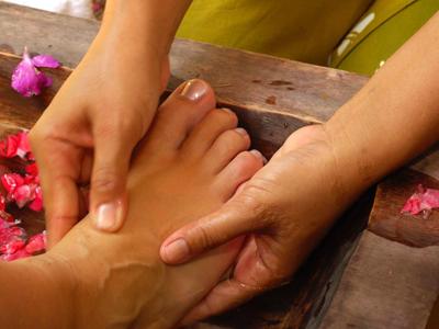 Eine Fußmassage mit Händen, Holzwanne und roten Blüten auf braunem Untergrund.