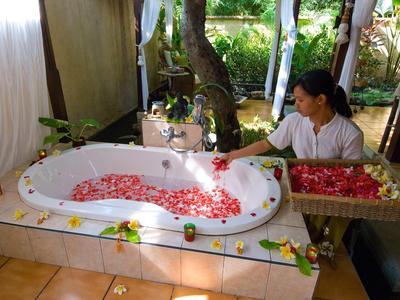 Frau dekoriert mit Rosenblättern eine weiße Herz-Badewanne in tropischer, offener Umgebung.
