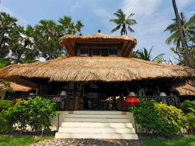 Ein großes, offenes Haus mit Strohdach, umgeben von grünen Pflanzen und Palmen im Hintergrund.