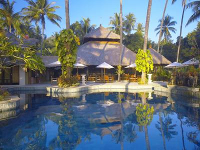 Ein ruhiger Pool spiegelt Palmen und ein rundes, strohgedecktes Gebäude in tropischer Umgebung.