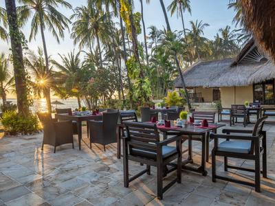 Terrassentisch mit Stühlen aus Holz bei Sonnenuntergang, umgeben von Palmen und einem strohgedeckten Dach.