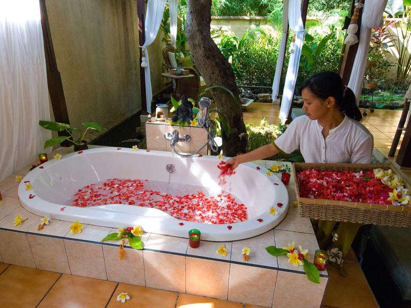 Frau dekoriert mit Rosenblättern eine weiße Herz-Badewanne in tropischer, offener Umgebung.