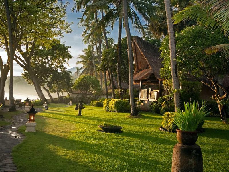 Grüner Garten mit Palmen, traditionellen Hütten und Sonnenlicht am Seeufer.