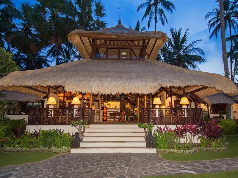 Großes strohgedecktes Haus mit warmem Licht, Palmen im Hintergrund und Treppe im Vordergrund.