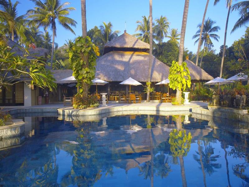 Ein ruhiger Pool spiegelt Palmen und ein rundes, strohgedecktes Gebäude in tropischer Umgebung.
