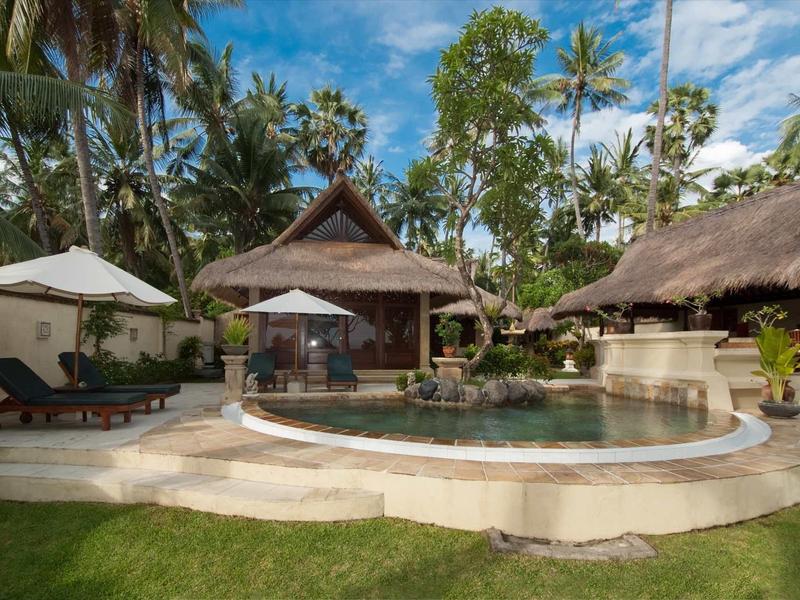 Runder Pool mit Liegestühlen, Palmen und traditionellen Holzhütten unter blauem Himmel.