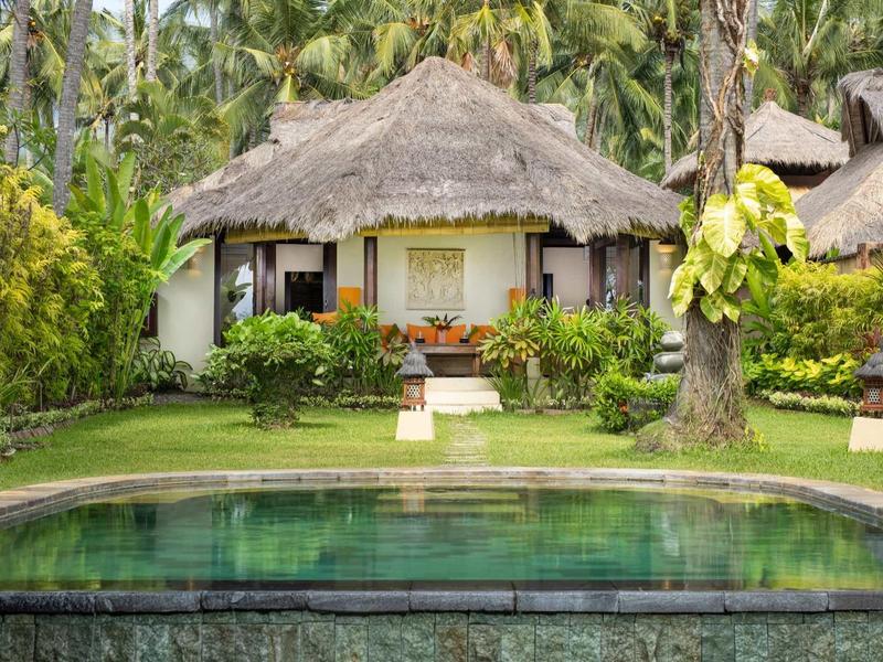 Ein strohgedecktes Haus mit Veranda, umgeben von grünem Gras und Palmen, vor einem Teich.