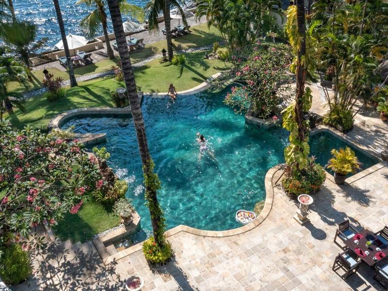 Blick auf grünen Pool mit Palmen, Liegestühlen und Personen, umgeben von Terrasse und Garten.