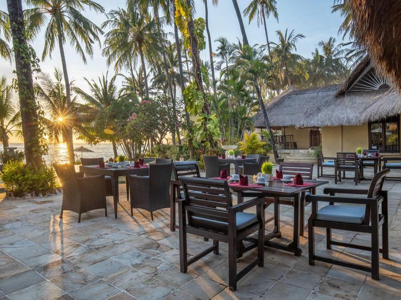 Terrassentisch mit Stühlen aus Holz bei Sonnenuntergang, umgeben von Palmen und einem strohgedeckten Dach.