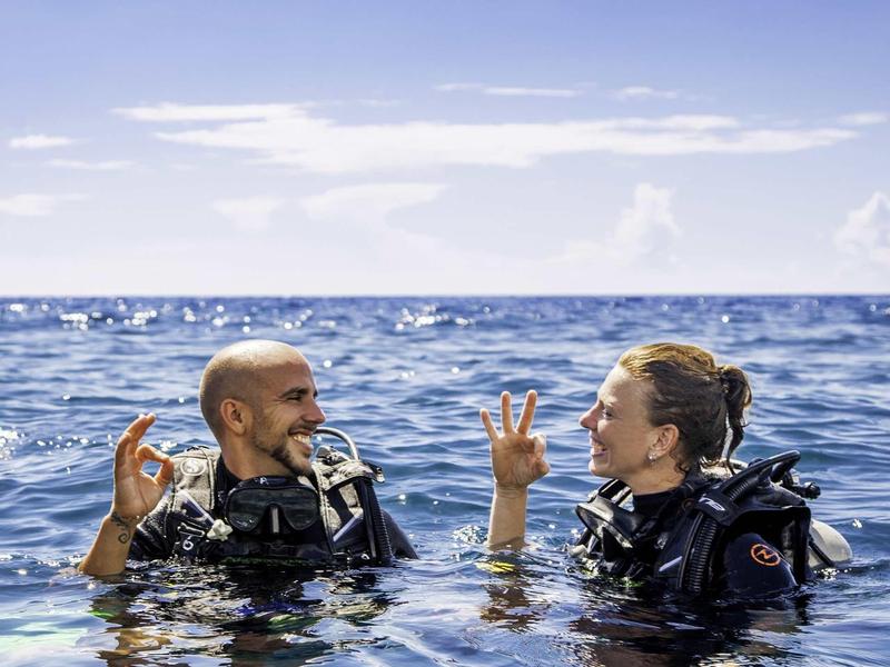 Zwei Taucher im blauen Meer zeigen sich mit Händen das OK-Zeichen bei klarem Himmel.