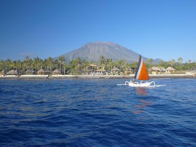Żaglówka z pomarańczowym żaglem na niebieskim morzu przed zalesionym wybrzeżem i górami.