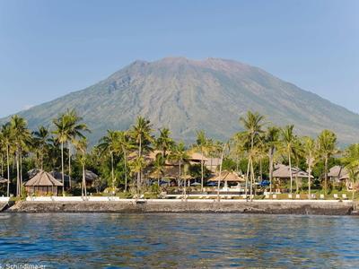 Luksusowe wille kurortu nad brzegiem z palmami przed dużym wulkanem w tle.