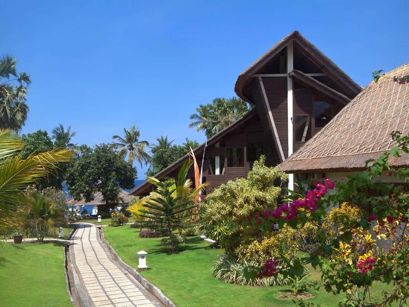 Ścieżka przez tropikalny ogród prowadząca do tradycyjnych budynków z dachami ze słomy pod jasnym niebem.