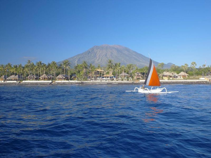 Żaglówka z pomarańczowym żaglem na niebieskim morzu przed zalesionym wybrzeżem i górami.