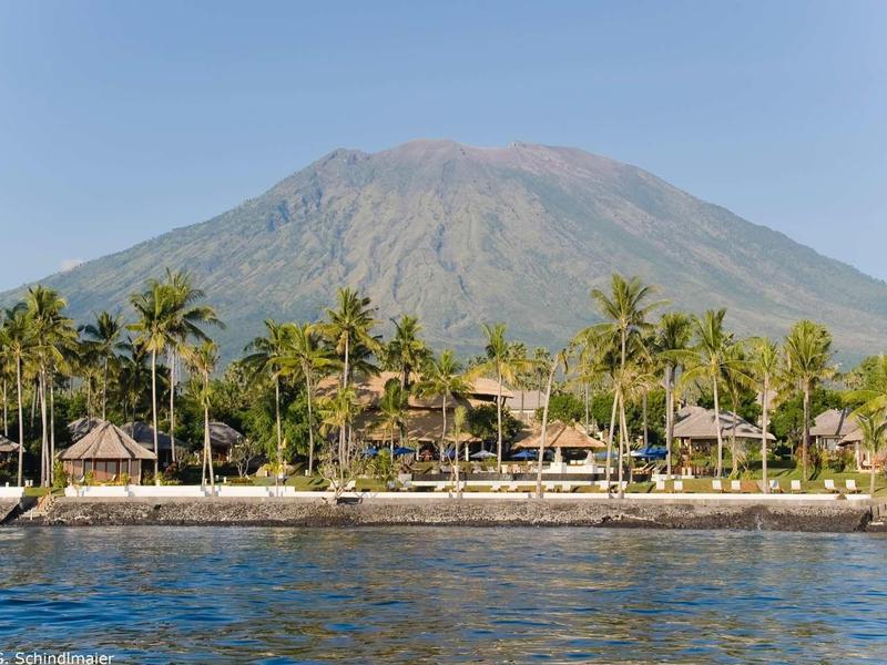 Luksusowe wille kurortu nad brzegiem z palmami przed dużym wulkanem w tle.