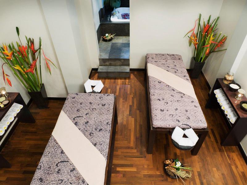 Zwei Massageliegen mit grauen Decken, Holzfußboden und Blumenarrangements in einem Behandlungsraum.