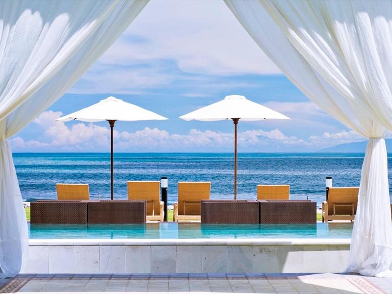 Meerblick mit Pool, weißen Vorhängen, Sonnenschirmen und Liegestühlen unter blauem Himmel.