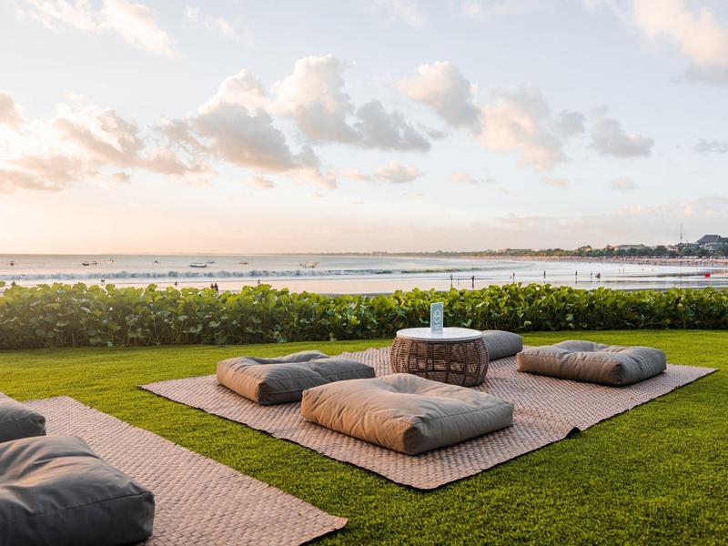 Outdoor-Sitzbereich mit Kissen und Teppichen auf grünem Rasen, Blick auf ruhiges Wasser und Himmel.