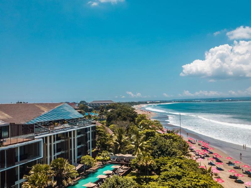 Strandresort met zwembad, palmbomen en veel parasols langs de kust bij een blauwe lucht.