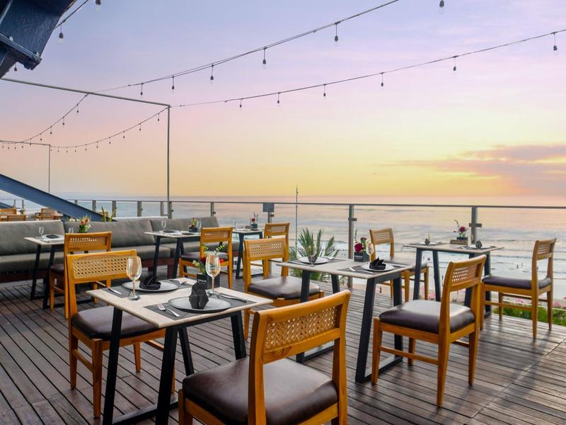 Terras van een restaurant met houten tafels en stoelen met uitzicht op het strand bij zonsondergang.