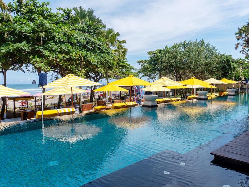 Groot hotelzwembad met gele parasols en ligstoelen naast groene bomen.