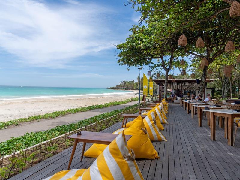 Lang houten dek met stoelen en gele kussens op het strand onder bomen.