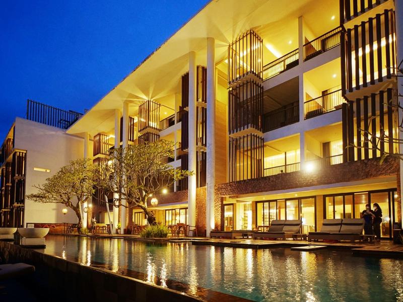 Modern hotelgebouw met balkons, verlicht in de avond, met een groot zwembad op de voorgrond.