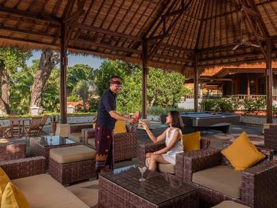 Salon extérieur d'hôtel avec meubles en osier et serveur donnant un verre à un client sous un toit de chaume.