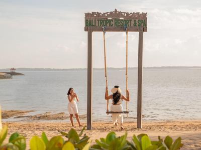 Couple profitant d'une balançoire au bord de la mer au coucher du soleil avec verdure tropicale au premier plan.