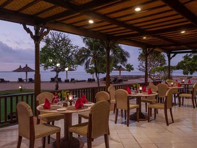 Espace repas en plein air au bord de la mer avec des tables dressées sous une terrasse couverte au coucher du soleil.