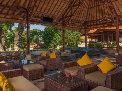 Überdachte Lounge mit Rattanmöbeln, gelben Kissen und Blick auf grüne Bäume bei Tageslicht.