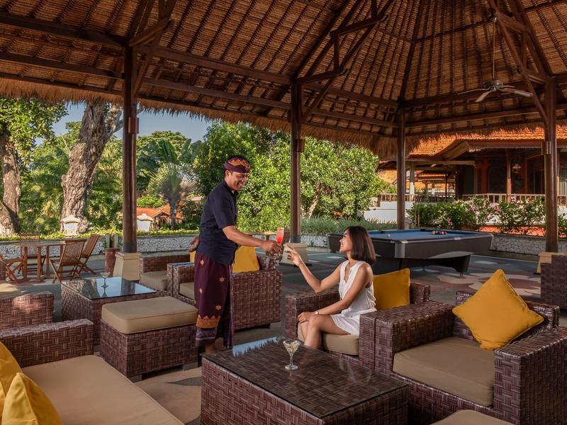 Salon extérieur d'hôtel avec meubles en osier et serveur donnant un verre à un client sous un toit de chaume.