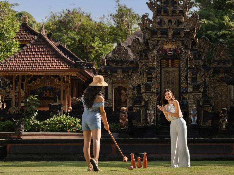 Deux femmes jouent avec des maillets en bois dans un jardin tropical près d'une architecture balinaise traditionnelle.