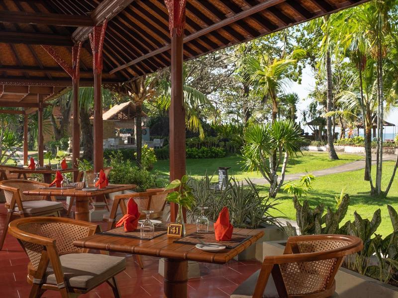 Espace repas extérieur avec tables et chaises en bois donnant sur un jardin luxuriant et des palmiers.