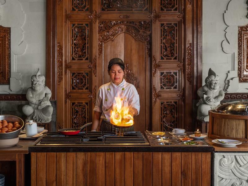 Chef cuisinier préparant un plat flambé dans une cuisine élégante avec décor en bois et sculptures traditionnelles.