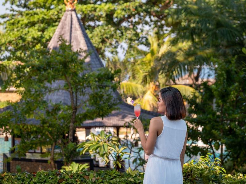 Femme en robe blanche prenant une photo près d'une végétation luxuriante et d'une structure au toit pointu.