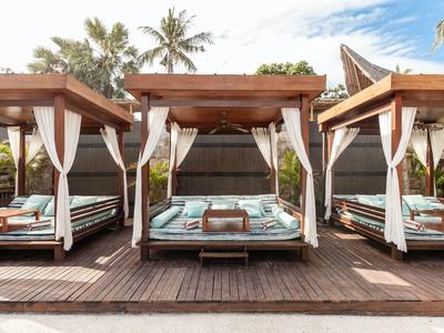 Trzy drewniane łóżka plażowe z białymi zasłonami i turkusowymi poduszkami na tarasie pod palmami.