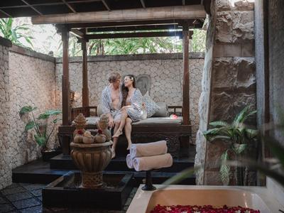 Para odpoczywa na zadaszonej drewnianej ławce w ogrodzie z roślinami i kamiennym murem.