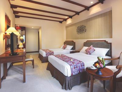 Pokój hotelowy z dwoma łóżkami, biurkiem i wygodnym fotelem w ciepłych kolorach.