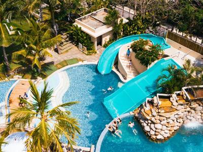 Basen na zewnątrz ze zjeżdżalnią wodną, otoczony palmami i leżakami w tropikalnym kurorcie.