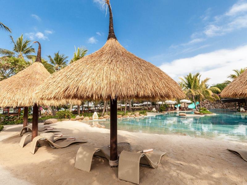 Plaża z parasolami z trzciny i leżakami obok basenu pod niebieskim niebem.