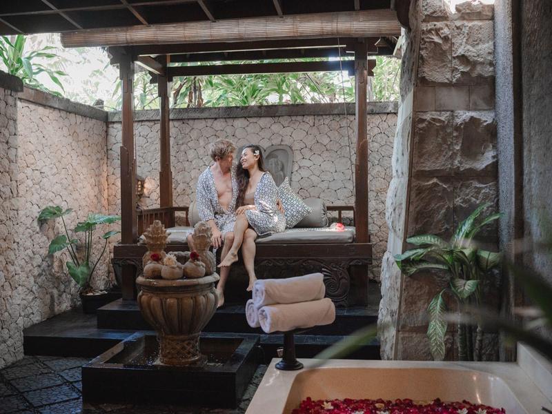 Para odpoczywa na zadaszonej drewnianej ławce w ogrodzie z roślinami i kamiennym murem.