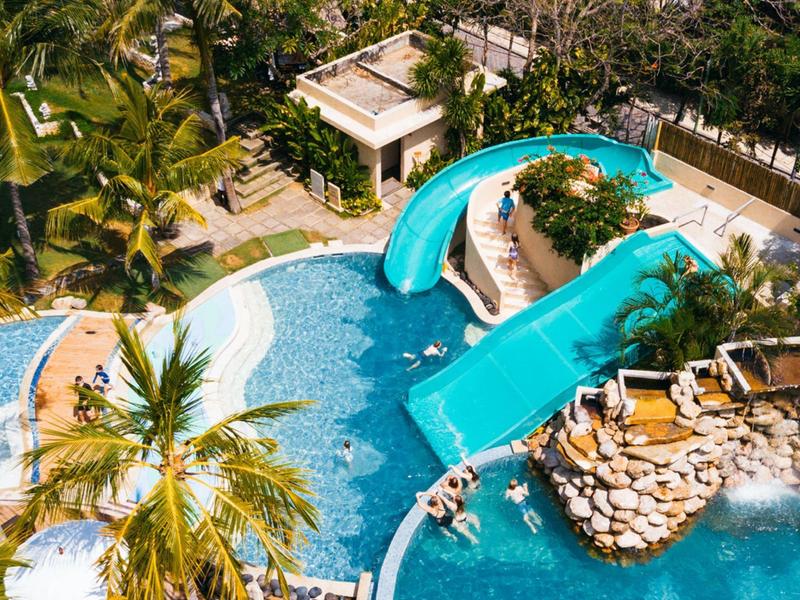 Basen na zewnątrz ze zjeżdżalnią wodną, otoczony palmami i leżakami w tropikalnym kurorcie.