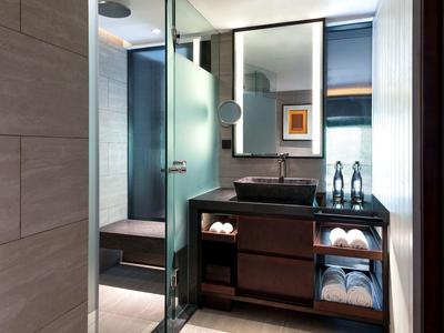 Moderní hotelová koupelna se skleněnou sprchou, tmavou umyvadlem a ručníky na otevřených policích.