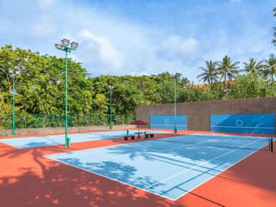 Campo sportivo all'aperto con canestro da basket e fari, circondato da alberi e palme.