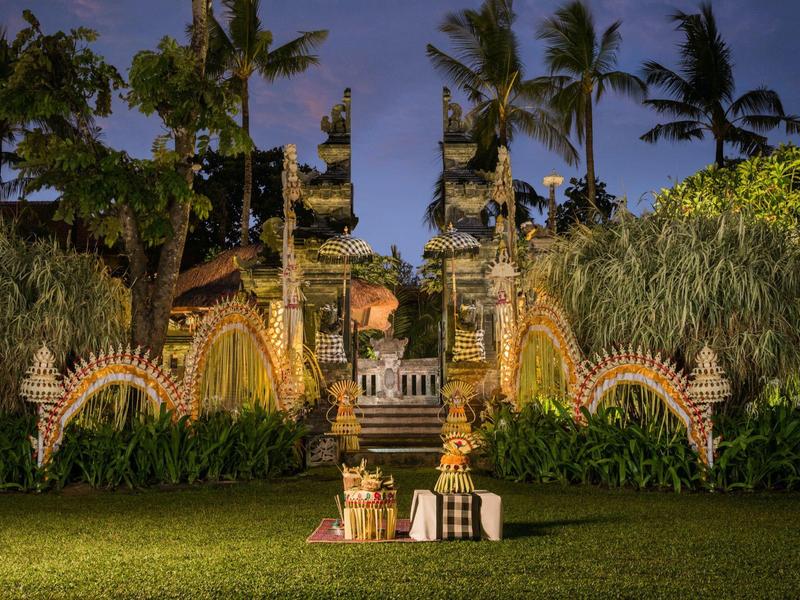 Večerní pohled na bohatě zdobenou tropickou zahradu s palmami a sochami.